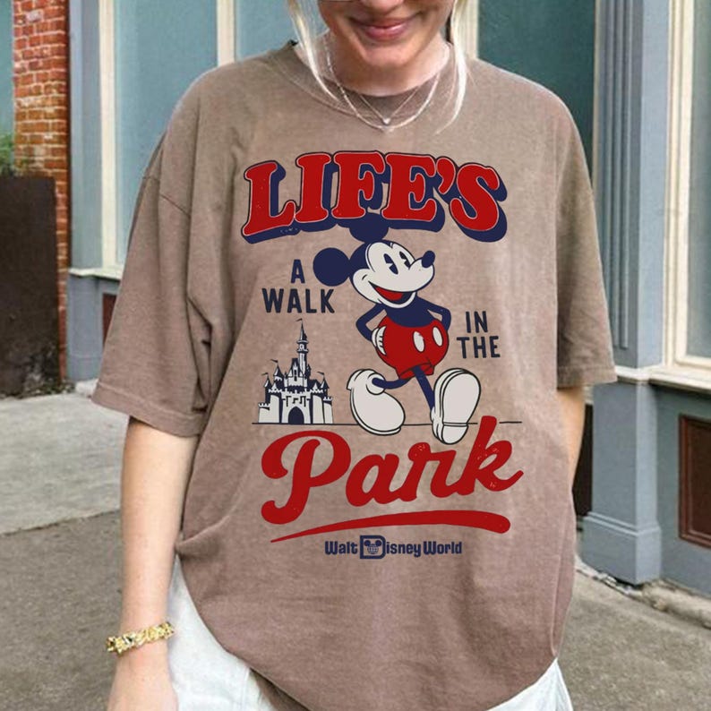 Puede incluir: Camiseta marr&oacute;n con Mickey Mouse y el texto "LIFE'S A WALK IN THE PARK" en rojo y azul. El dise&ntilde;o incluye un castillo y las palabras "Walt Disney World".