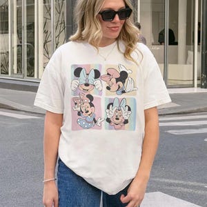 May include: Cream-colored t-shirt featuring a four-panel design of Minnie Mouse. Each panel showcases a different pose with a pastel rainbow background. The shirt is worn by a person wearing sunglasses and blue jeans.