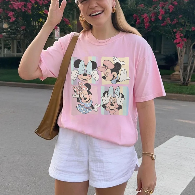 May include: A light pink t-shirt featuring four images of Minnie Mouse in different poses. The shirt is paired with white shorts and a brown shoulder bag. The overall style is casual and summery.