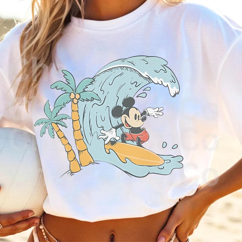 May include: White t-shirt featuring a cartoon Mickey Mouse surfing a wave. The design includes palm trees and a yellow surfboard. The wave is light blue, and Mickey is wearing red shorts. A fun, summery graphic tee.