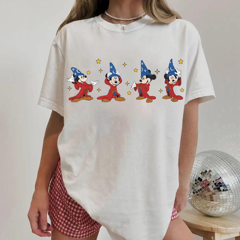 Peut inclure: T-shirt blanc avec un dessin anim&eacute; de Mickey Mouse en costume de magicien. Mickey est repr&eacute;sent&eacute; avec une robe rouge et un chapeau de magicien bleu, avec des &eacute;toiles jaunes. Le t-shirt est de style d&eacute;contract&eacute; et unisexe.