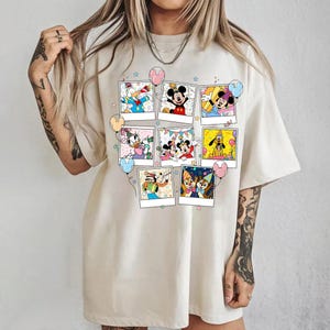 Puede incluir: Camiseta color crema con un collage de im&aacute;genes de personajes de dibujos animados en marcos estilo polaroid. Las im&aacute;genes incluyen Mickey Mouse, Minnie Mouse, Donald Duck y Pluto. Globos y estrellas decoran el dise&ntilde;o.