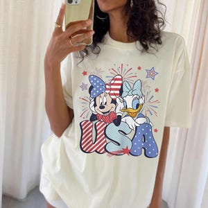 May include: Cream-colored t-shirt featuring Minnie Mouse and Daisy Duck with patriotic bows, fireworks, and the word "USA" in red, white, and blue. The graphic tee is a casual, comfortable style.