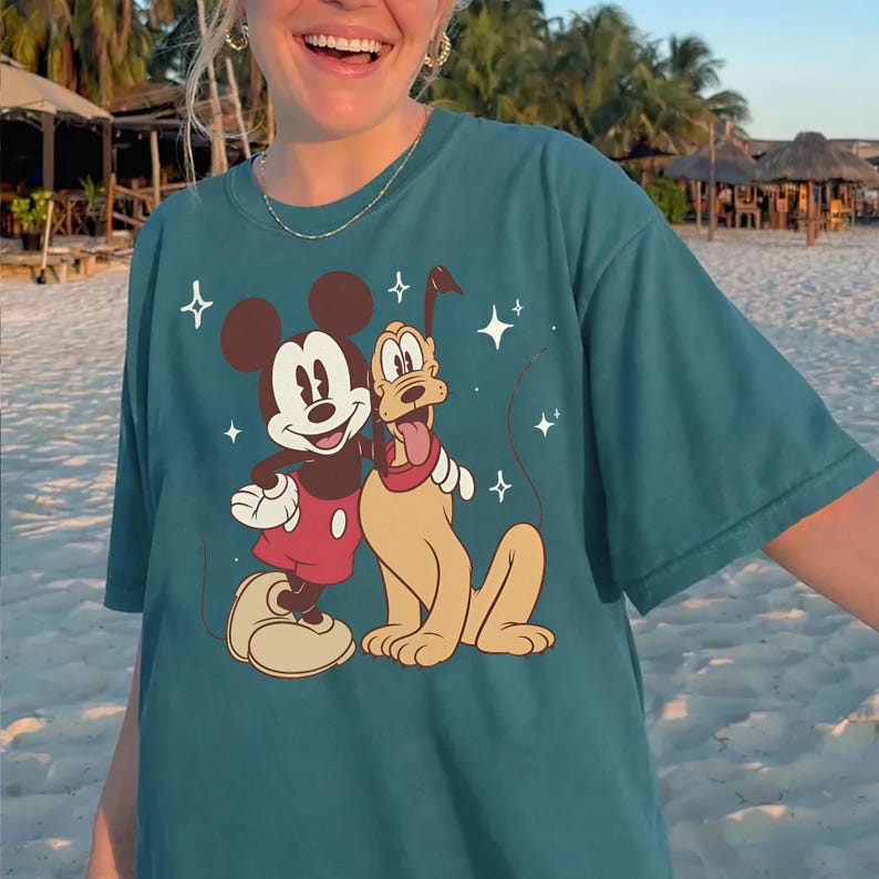 May include: Teal t-shirt with a graphic of Mickey Mouse and Pluto. Mickey wears red shorts, and Pluto has a red collar. The design includes white stars. The tee is made of a soft, comfortable material.