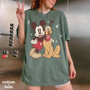 May include: Green t-shirt featuring Mickey Mouse and Pluto cartoon graphic. The graphic shows Mickey Mouse in a red shirt and Pluto with a red collar. The shirt has the text "cotton + luxe" at the bottom.