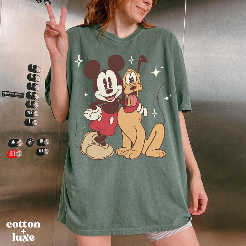 May include: Green t-shirt featuring Mickey Mouse and Pluto cartoon graphic. The graphic shows Mickey Mouse in a red shirt and Pluto with a red collar. The shirt has the text "cotton + luxe" at the bottom.