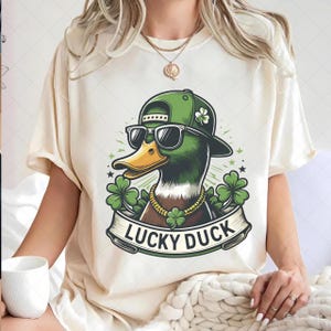 Pode incluir: Camiseta creme com um pato de desenho animado usando óculos de sol, boné verde e corrente dourada. O pato é cercado por trevos e a frase "LUCKY DUCK" em uma faixa.