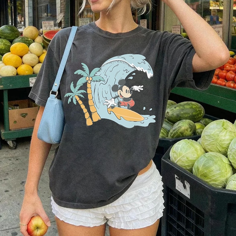 May include: Dark gray t-shirt featuring a graphic of Mickey Mouse surfing a wave with palm trees. The shirt is paired with white ruffled shorts and a light blue shoulder bag. The background shows a fruit stand.