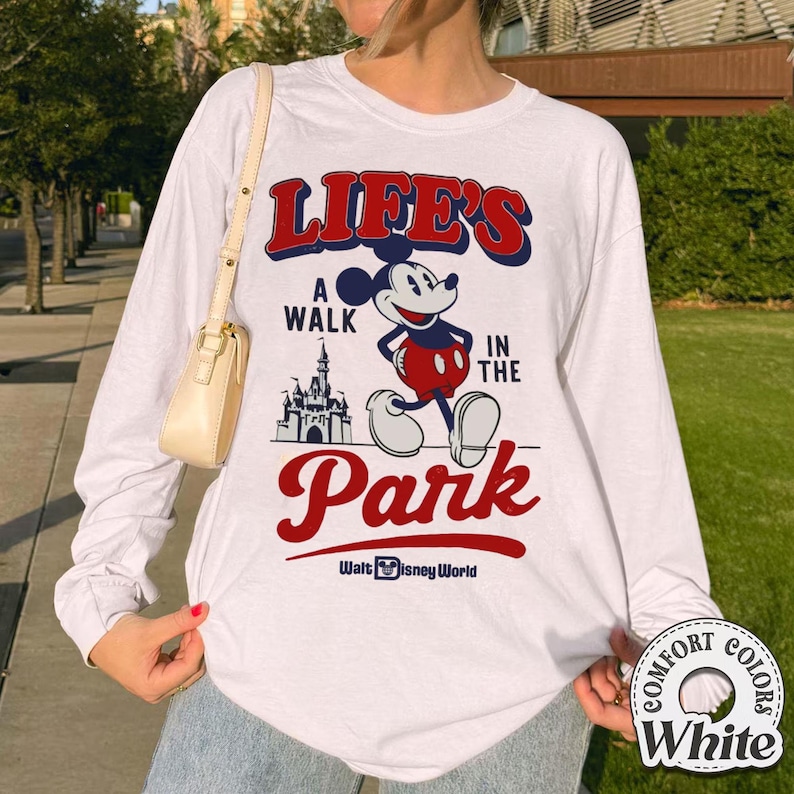 Puede incluir: Camiseta blanca de manga larga con un gr&aacute;fico de Mickey Mouse y el texto "LIFE'S A WALK IN THE PARK" en rojo y azul. La camiseta tambi&eacute;n incluye el logotipo de Walt Disney World.