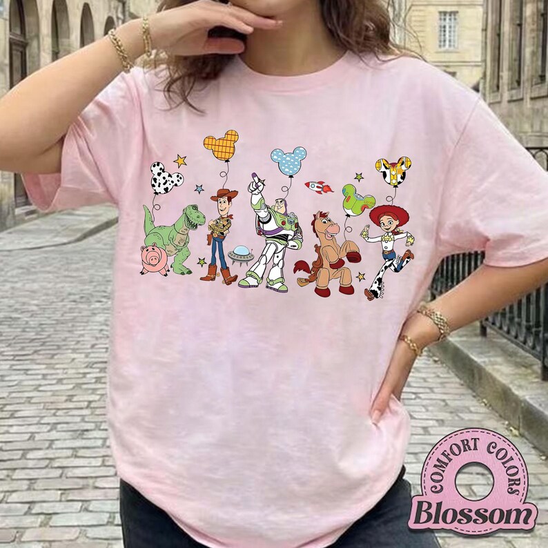 May include: Pink t-shirt featuring a colorful graphic of Toy Story characters and balloons. The design includes Woody, Buzz Lightyear, Jessie, and other characters. The shirt has the "Comfort Colors" and "Blossom" logo.