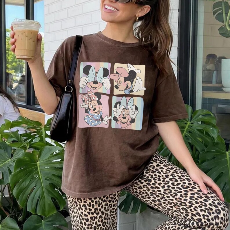 May include: Brown t-shirt featuring four panels with Minnie Mouse illustrations in pastel colors. The shirt is worn with leopard print pants. The person is holding a clear plastic cup with a brown beverage.