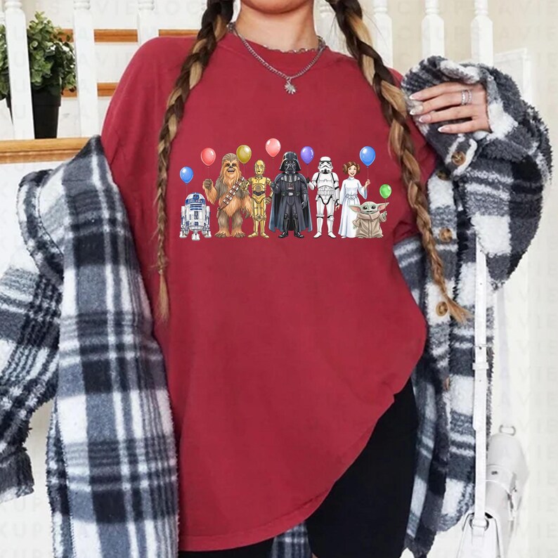 May include: A maroon t-shirt with a Star Wars character graphic. The design includes R2-D2, Chewbacca, C-3PO, Darth Vader, a Stormtrooper, Princess Leia, and Grogu, with balloons. The shirt is a casual, crew neck style, perfect for fans.