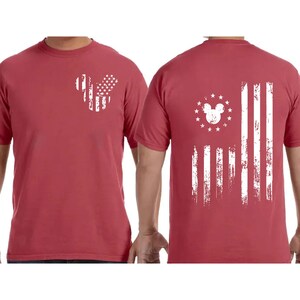 May include: Red t-shirt with a distressed white American flag design. The front features a Mickey Mouse head shape with stars and stripes. The back displays a larger flag design with a Mickey Mouse head and stars.