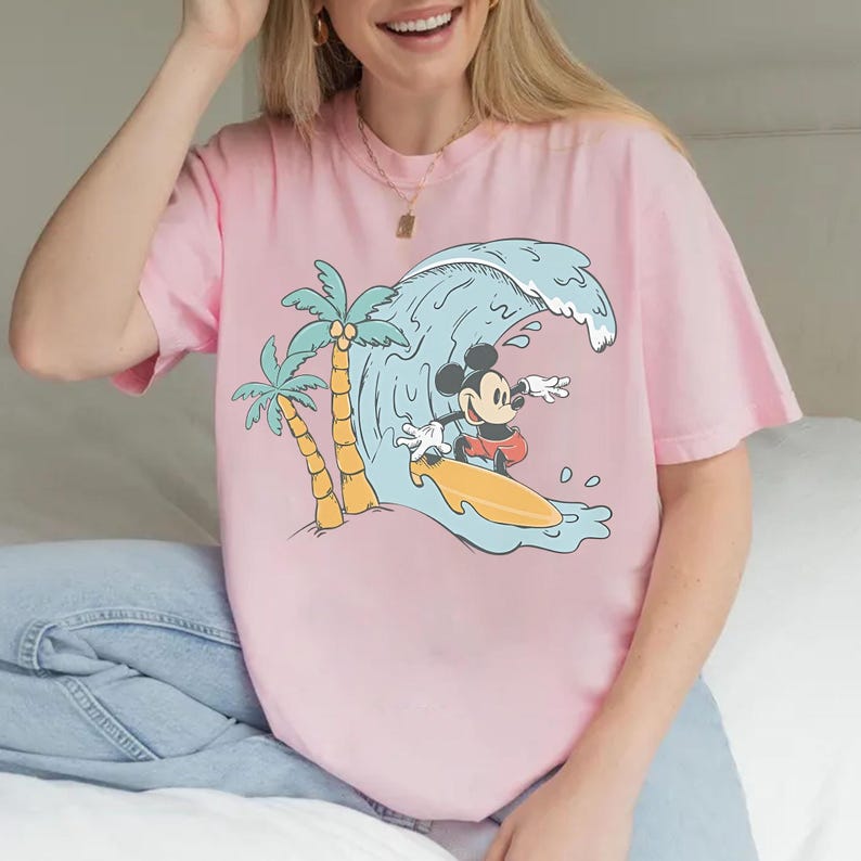 May include: Pink t-shirt featuring a graphic of Mickey Mouse surfing a wave. The design includes palm trees and a yellow surfboard. The shirt is a light pink color.