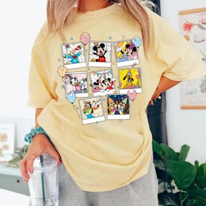 Puede incluir: Una camiseta amarilla clara con un collage de im&aacute;genes de personajes de dibujos animados en marcos estilo polaroid. Las im&aacute;genes incluyen Mickey Mouse, Donald Duck y Pluto. La camiseta tiene cuello redondo y mangas cortas.