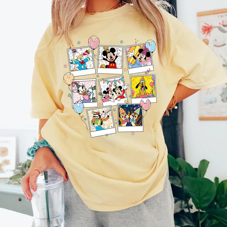 Puede incluir: Una camiseta amarilla clara con un collage de im&aacute;genes de personajes de dibujos animados en marcos estilo polaroid. Las im&aacute;genes incluyen Mickey Mouse, Donald Duck y Pluto. La camiseta tiene cuello redondo y mangas cortas.