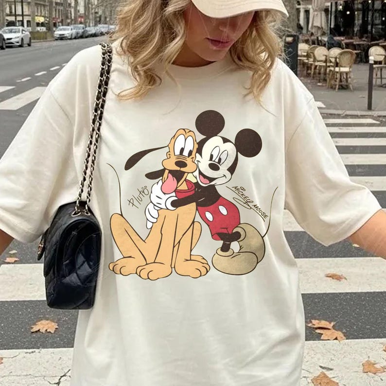 May include: Cream-colored t-shirt featuring a graphic of Mickey Mouse and Pluto embracing. Mickey Mouse is in his classic red shorts and Pluto is in a sitting position. The text "Mickey Mouse" and "Pluto" are also visible.