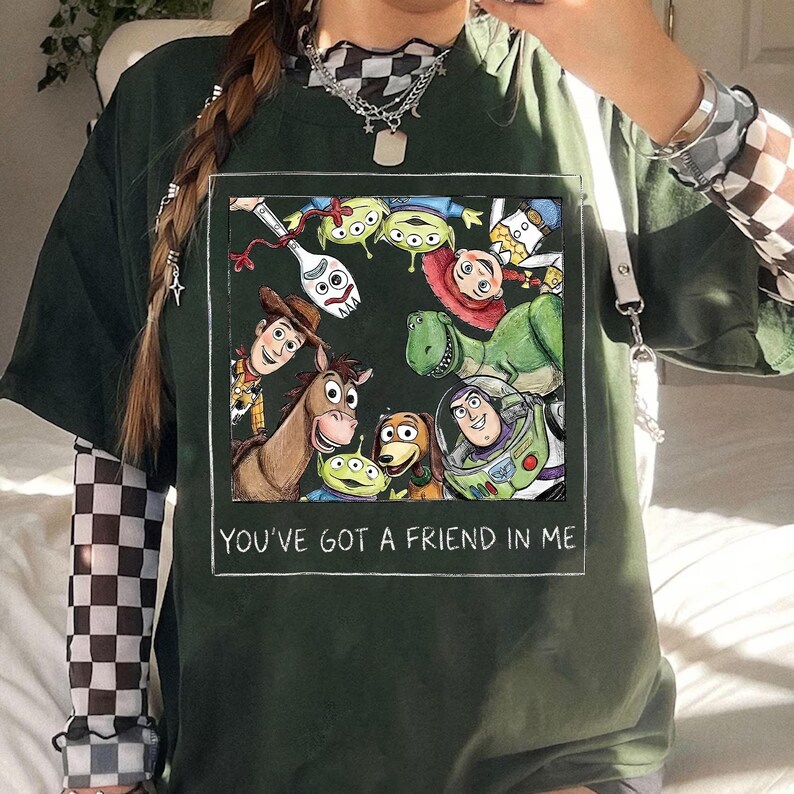 May include: Dark green t-shirt featuring a rectangular graphic of Toy Story characters. The graphic includes Woody, Buzz Lightyear, and other characters. The text "YOU'VE GOT A FRIEND IN ME" is written in white at the bottom of the graphic.