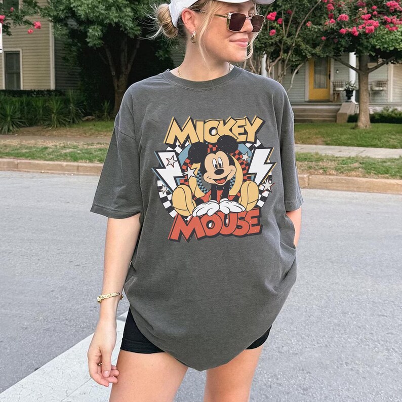 May include: Dark gray t-shirt featuring a colorful graphic of Mickey Mouse with the text "Mickey Mouse". The design includes lightning bolts, stars, and a checkered pattern. The shirt has short sleeves and a relaxed fit.