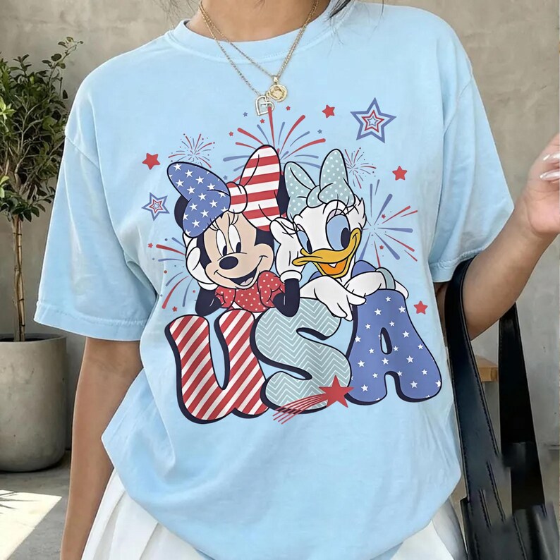 May include: Light blue t-shirt featuring Minnie Mouse and Daisy Duck with patriotic bows and the word "USA" in red, white, and blue. Fireworks and star graphics complete the design. A casual, festive top.