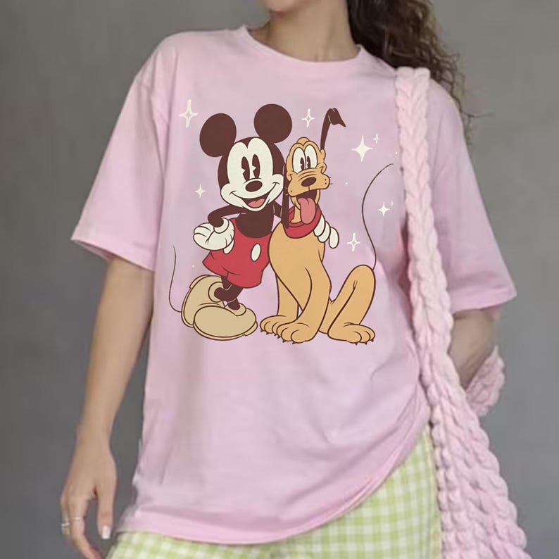 May include: A pale pink t-shirt with a graphic of Mickey Mouse and Pluto. Mickey is in red shorts and Pluto has a red collar. The design includes star accents. The shirt is a casual, short-sleeved style.