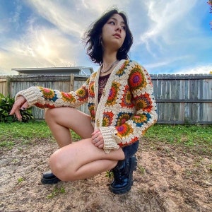 Puede incluir: Una mujer lleva una chaqueta de punto de ganchillo con un estampado floral colorido. La chaqueta es de color crema con una variedad de colores brillantes, incluyendo naranja, amarillo, verde y morado. La mujer lleva pantalones cortos negros y botas negras.