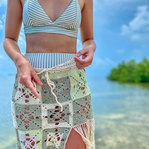 Puede incluir: Una mujer lleva un top de bikini a rayas verdes y blancas y una falda de ganchillo con flecos. La falda está hecha de cuadrados con un patrón floral. La mujer está de pie en una playa con agua en el fondo.