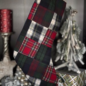 May include: A Christmas stocking with a patchwork design. The stocking features a mix of red, green, black, and white plaid fabric squares. The stocking is hanging in front of a dark background with Christmas decorations.