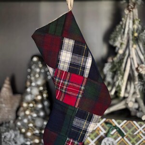 May include: A patchwork Christmas stocking in a plaid pattern. The stocking features a mix of red, green, navy, and white plaid fabrics. It is hanging from a decorative loop, with a blurred background of Christmas decorations.