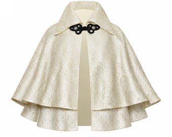 May include: Cream-colored cape with a floral pattern and a wide collar. The cape features a double-layered design and a black clasp closure at the neckline. The fabric has a subtle sheen, suggesting a formal or elegant style.