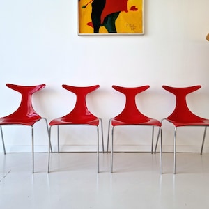 May include: Four modern red chairs with a unique, curved back design. The chairs have a glossy finish and are supported by slender, silver metal legs. The chairs are arranged in a row against a white wall.