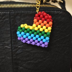 May include: A rainbow-colored beaded heart keychain with a gold-colored chain and clasp. The heart is made of small, round beads in a gradient of colors, including red, orange, yellow, green, blue, and purple. The keychain is attached to a black leather bag.