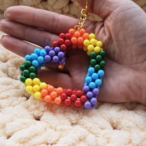 May include: A rainbow-colored beaded heart keychain with a gold-colored keyring. The heart is made of small, round beads in a spectrum of colors, including red, orange, yellow, green, blue, and purple. The keychain is held against a soft, cream-colored background.