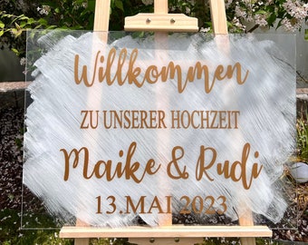 Op de afbeelding: Een wit acryl bord met een gouden penseelstreep ontwerp en de tekst "Willkommen zu unserer Hochzeit Maike & Rudi 13. Mai 2023". Het bord staat op een houten ezel.