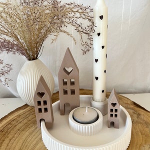 Può includere: Un portacandele in ceramica bianca con un bordo decorativo scanalato. Il portacandele presenta tre piccole case marroni con ritagli a forma di cuore e una candela bianca alta con cuori neri. Una piccola candela tealight si trova al centro del portacandele.