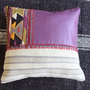 May include: A decorative pillow with a patchwork design. The pillow has a purple fabric center with a white and gray striped border. The pillow is decorated with colorful geometric patterns and red stitching.