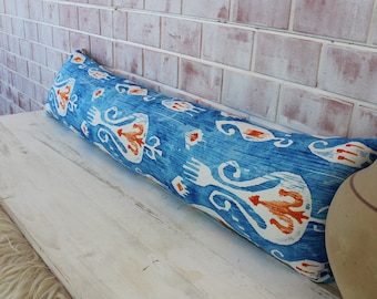 May include: A long, blue and white patterned bolster pillow with orange accents. The pillow is lying on a white wooden bench.