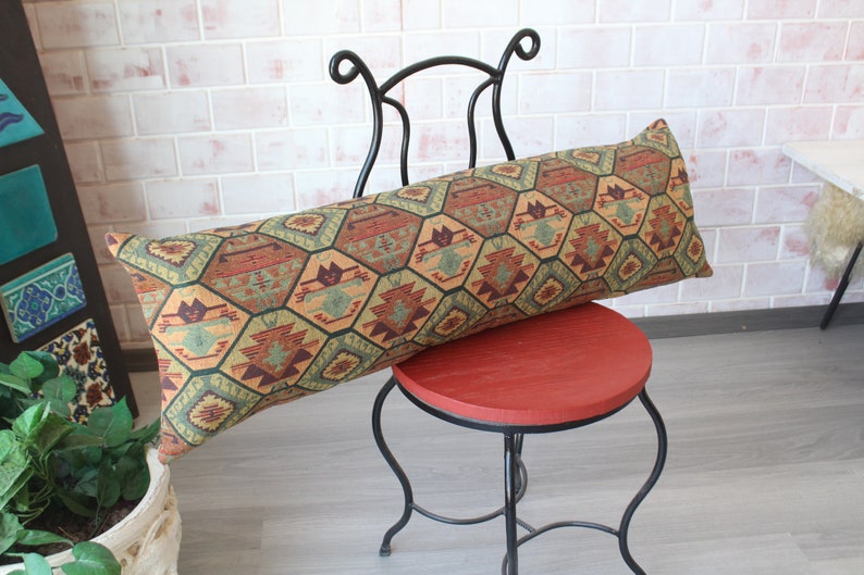 May include: A long, rectangular pillow with a geometric pattern in shades of green, brown, and orange. The pillow is resting on a black metal chair with a red seat.
