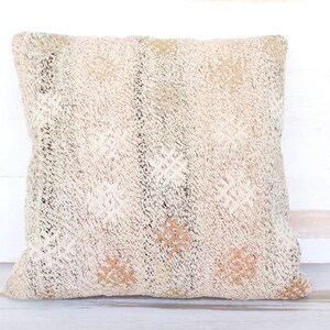 May include: Square decorative pillow with a textured, off-white surface. The pillow features a woven pattern with accents of brown, tan, and olive green. The design includes diamond and floral-like shapes, creating a rustic, bohemian aesthetic.