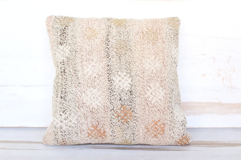 May include: Square decorative pillow with a textured, off-white surface. The pillow features a woven pattern with accents of brown, tan, and olive green. The design includes diamond and floral-like shapes, creating a rustic, bohemian aesthetic.