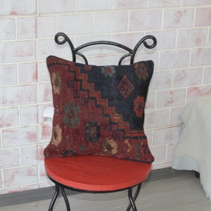 Puede incluir: Un coj&iacute;n de tiro rojo, negro y verde con dise&ntilde;os intrincados se sienta en un taburete de metal rojo. El coj&iacute;n est&aacute; hecho de una tela suave y tejida y tiene un aspecto vintage &uacute;nico.