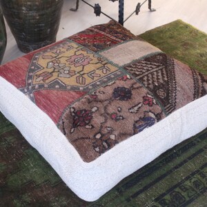 May include: Square ottoman with a patchwork design. The top features a mix of red, brown, and gold patterned fabric. The sides are a solid white fabric. The ottoman is placed on a green rug.