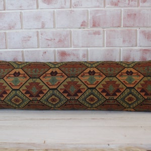 May include: A long, rectangular pillow with a geometric pattern in shades of brown, green, and orange. The pillow has a woven texture and is made of fabric.