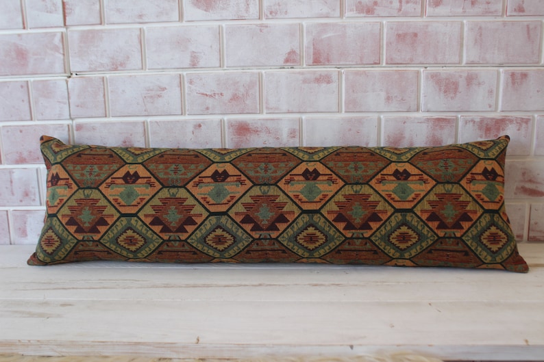 May include: A long, rectangular pillow with a geometric pattern in shades of brown, green, and orange. The pillow has a woven texture and is made of fabric.