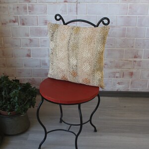 May include: A decorative pillow with a beige and gold pattern rests on a small, round, red chair. The chair has a black, wrought-iron frame. The background features a brick wall and decorative tiles.