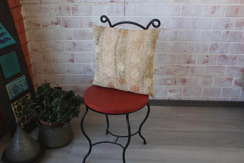 May include: A decorative pillow with a beige and gold pattern rests on a small, round, red chair. The chair has a black, wrought-iron frame. The background features a brick wall and decorative tiles.