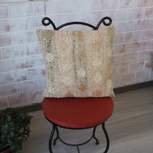 May include: A decorative pillow with a beige and gold patterned design rests on a small, round, red stool with black metal legs. The pillow has a textured appearance and is square-shaped. The background features a brick wall.