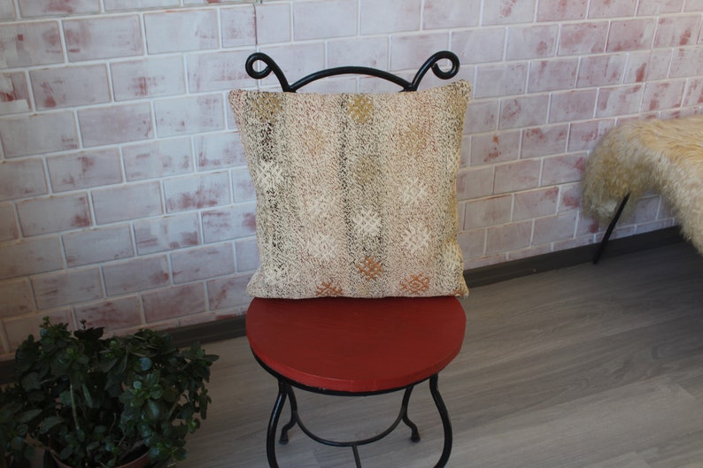 May include: A decorative pillow with a beige and gold patterned design rests on a small, round, red stool with black metal legs. The pillow has a textured appearance and is square-shaped. The background features a brick wall.