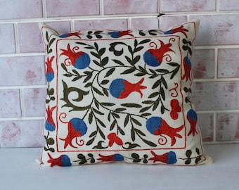 May include: A white pillow with a red, blue, and green embroidered design of pomegranates and leaves.