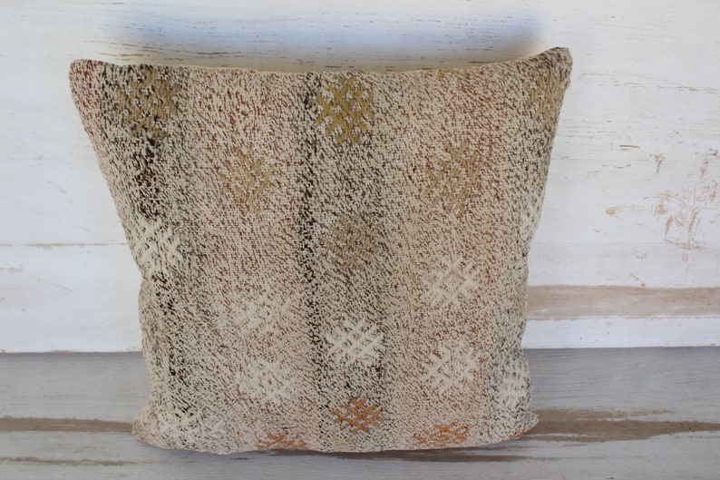 May include: A square, textured pillow with a neutral color palette. The pillow features a woven design with cream, brown, and tan hues. The pillow's surface has a raised, tactile quality, adding visual interest and depth. This decorative pillow is suitable for home decor.